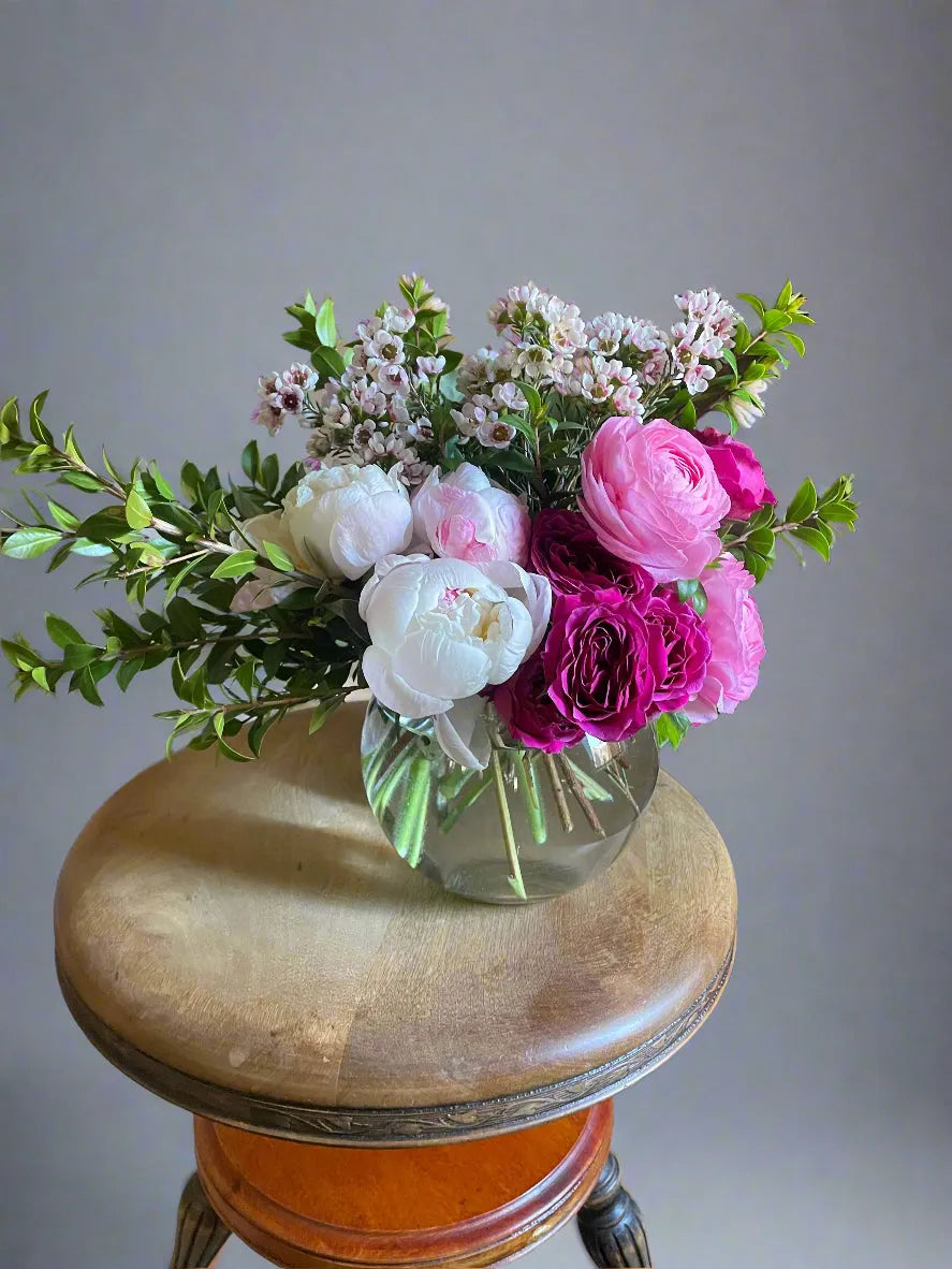 Bright pink and white peonies with greenery in a round glass vase on a wooden stool