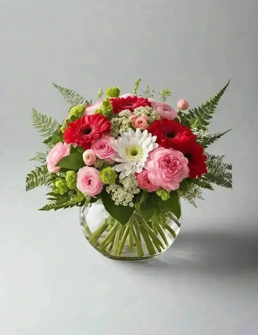 Bright fresh flower arrangement with pink roses, red gerbera daisies, white daisy, and green ferns in glass vase