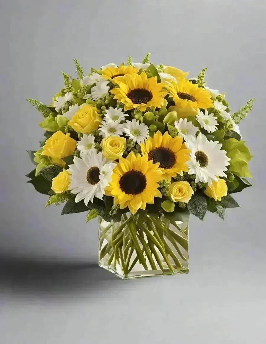 Bright yellow sunflowers and roses with white daisies in a clear glass vase bouquet
