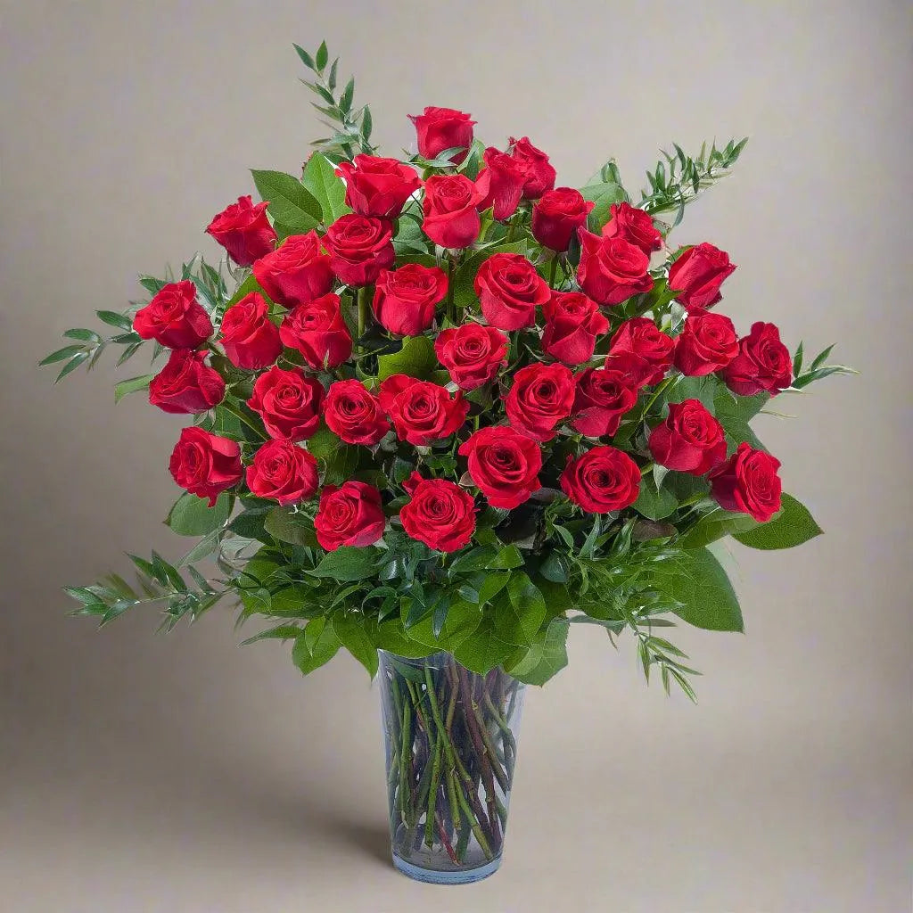 A three dozen long-stemmed red roses arranged in a clear vase, with green leaves and stems visible.