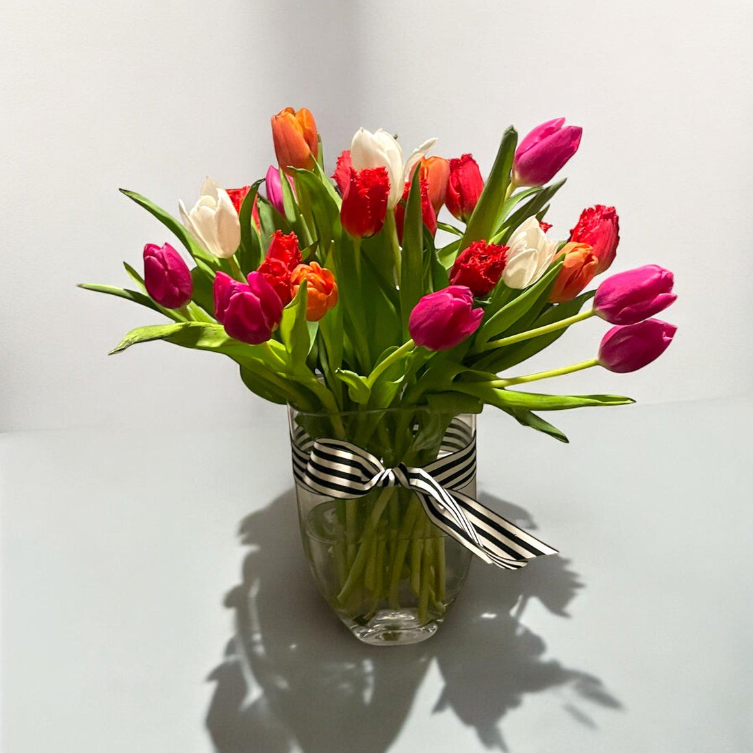 Bright bouquet of pink, red, orange, and white tulips in clear vase with black and white ribbon