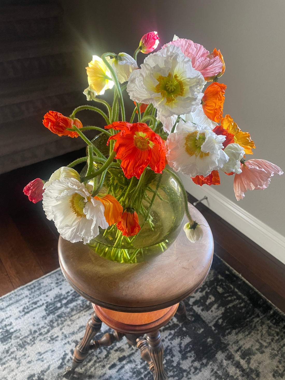 Bright bouquet of white, orange, and pink poppies in a round glass vase on a wooden stool