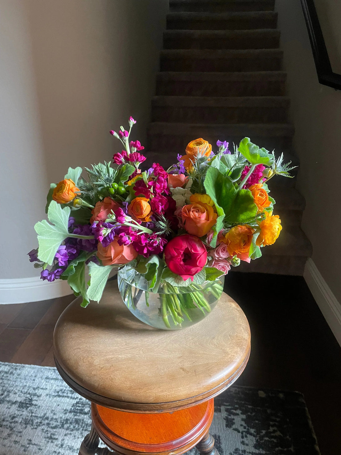 Bright mixed flower bouquet in clear glass vase on wooden stool by carpeted stairs