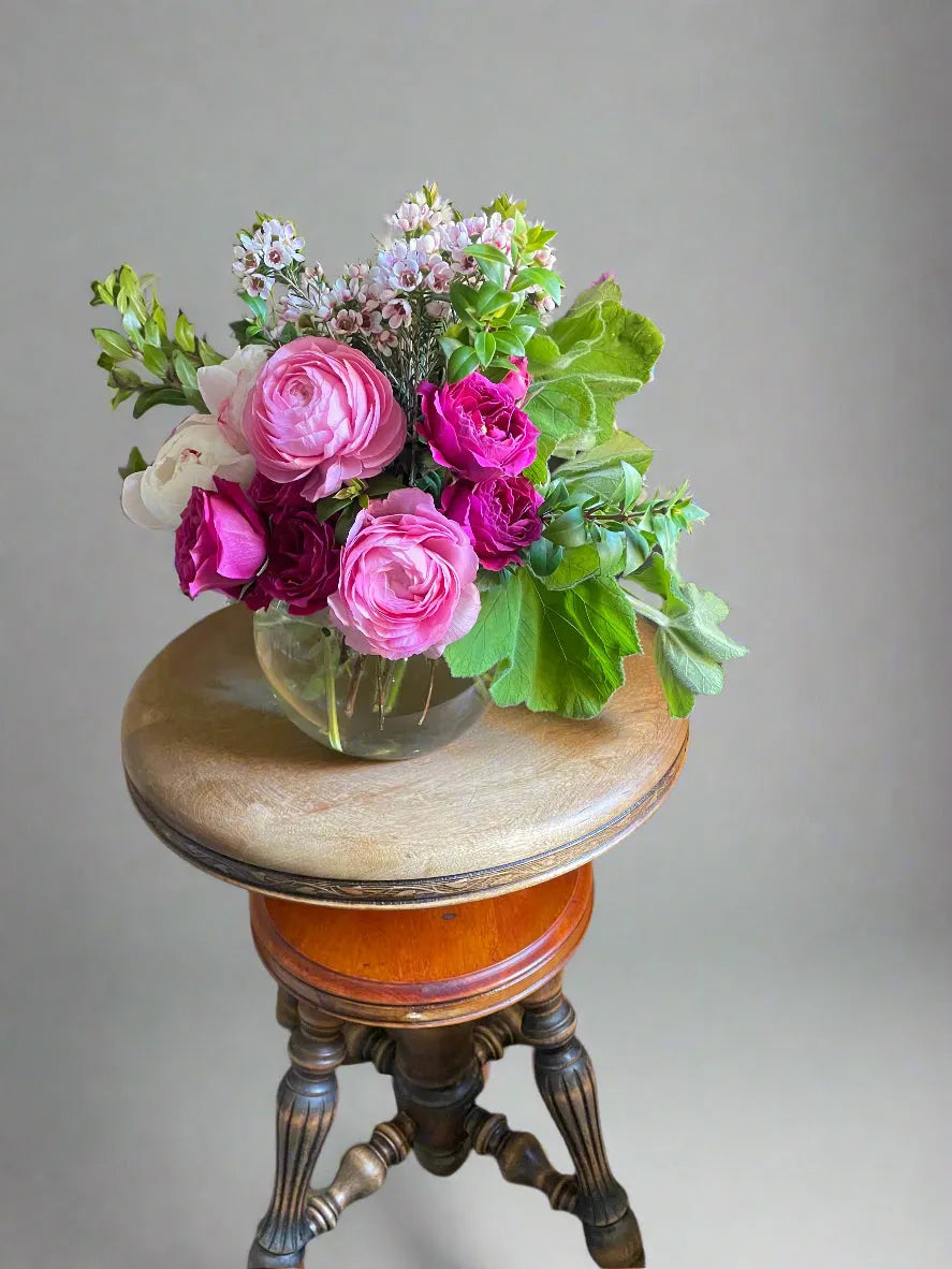 A stunning flower arrangement in a bubble bowl with pink ranunculus, white peonies, fusca spray roses, white wax flowers, and geranium greens.