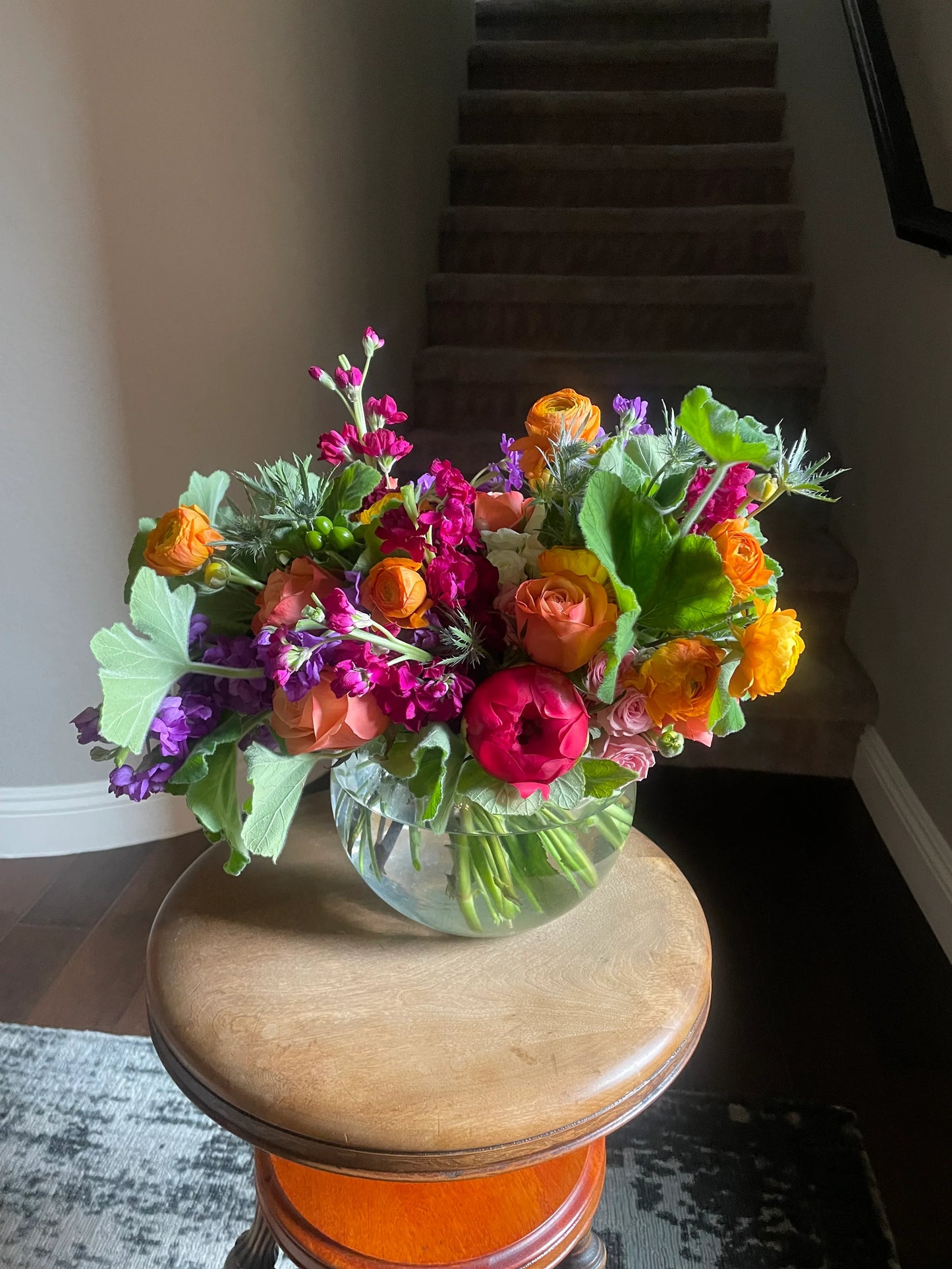 A stunning, colorful flower arrangement in a bubble container with roses, ranunculus, peonies, hypericum berries, and stock.