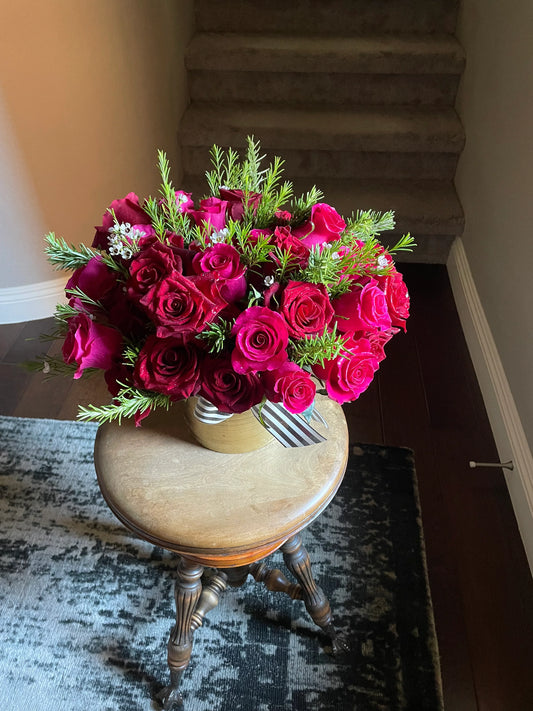 Red and fuschia roses with rosemary and wax flower accents in a gold vase with a black and white stripped ribbon.