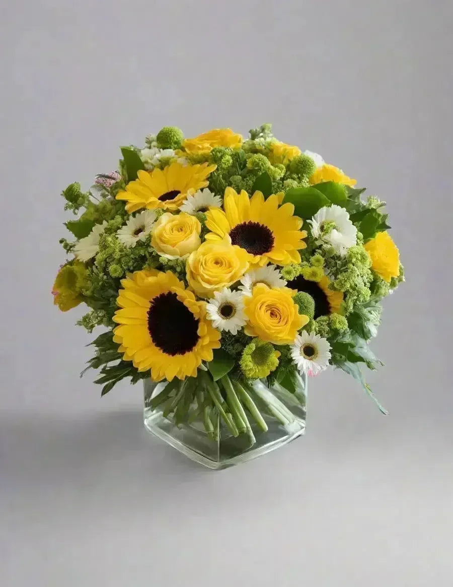 Bouquet of sunflowers, roses, and daisies in a clear vase on a light gray background